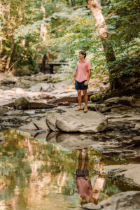 Walton high school senior standing on a rock by a creek with his reflection in the water during senior photos