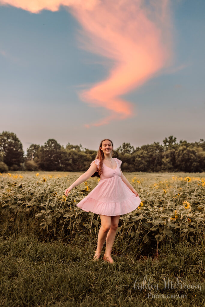 High school senior in a pink dress dancing beneath a beautiful pink cloud