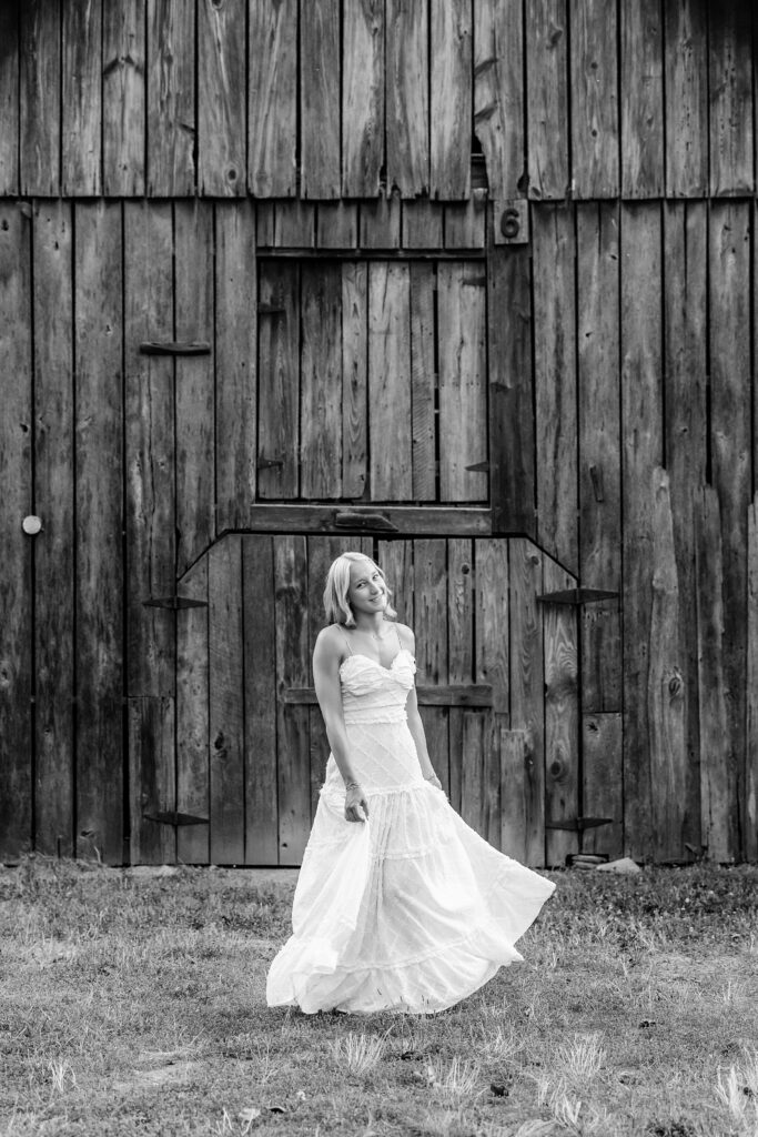 Black and white full-length senior portrait in a white dress in front of a rustic wooden barn